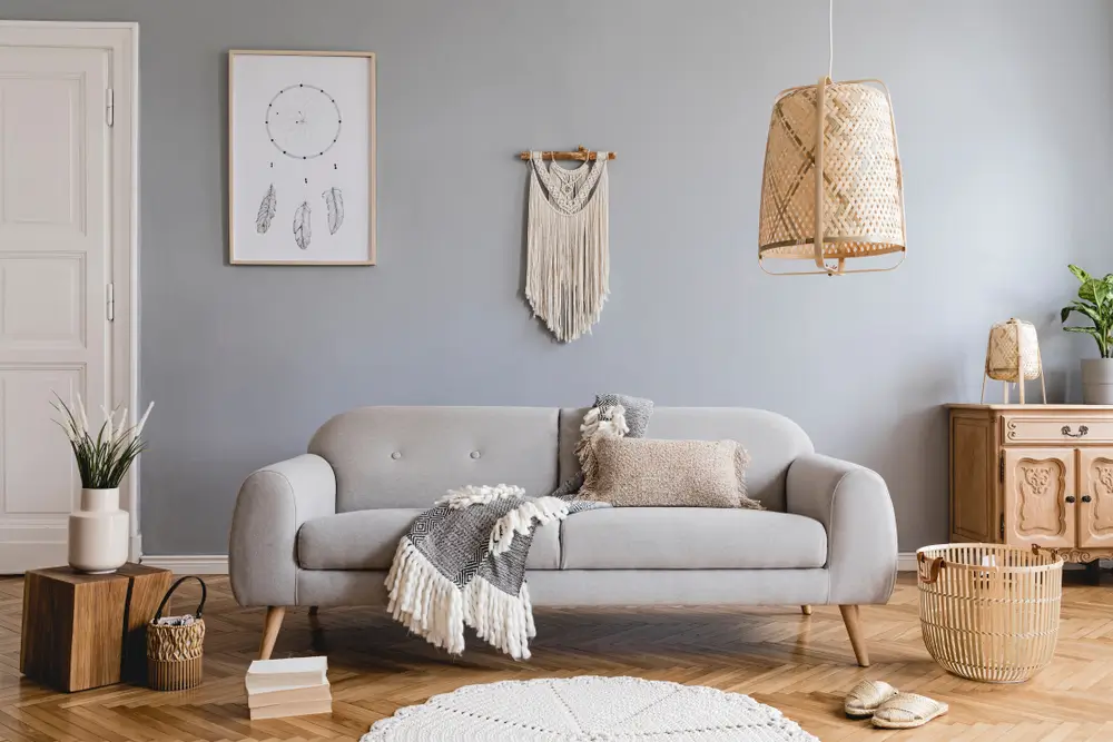 Composition bohème pour un intérieur de salon avec canapé gris, cube en bois, commode, macramé beige, lampe en rotin, plantes, plaid, fleurs et accessoires élégants.
